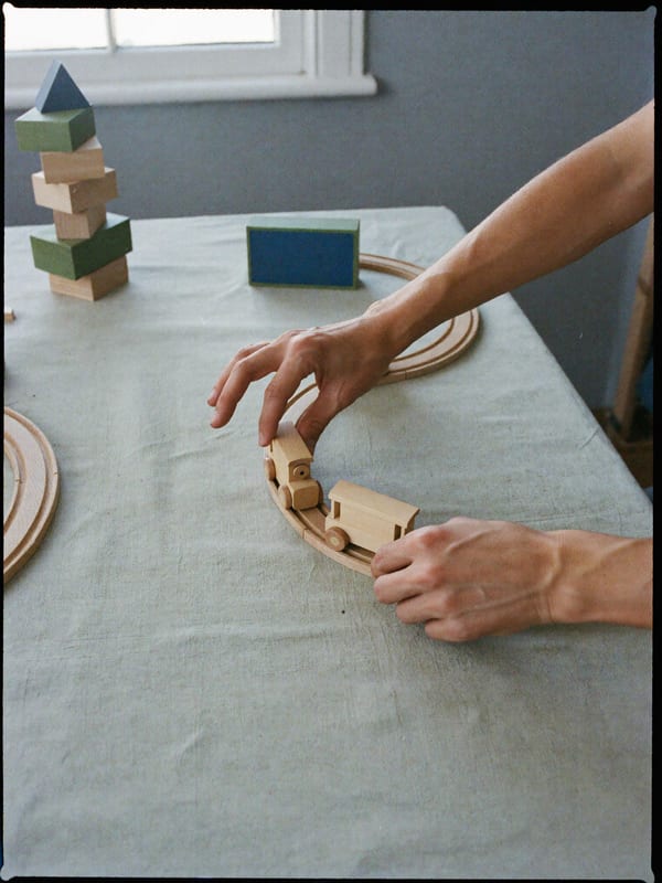 Two hands set a beech wood toy train on a wooden track beside stacking blocks